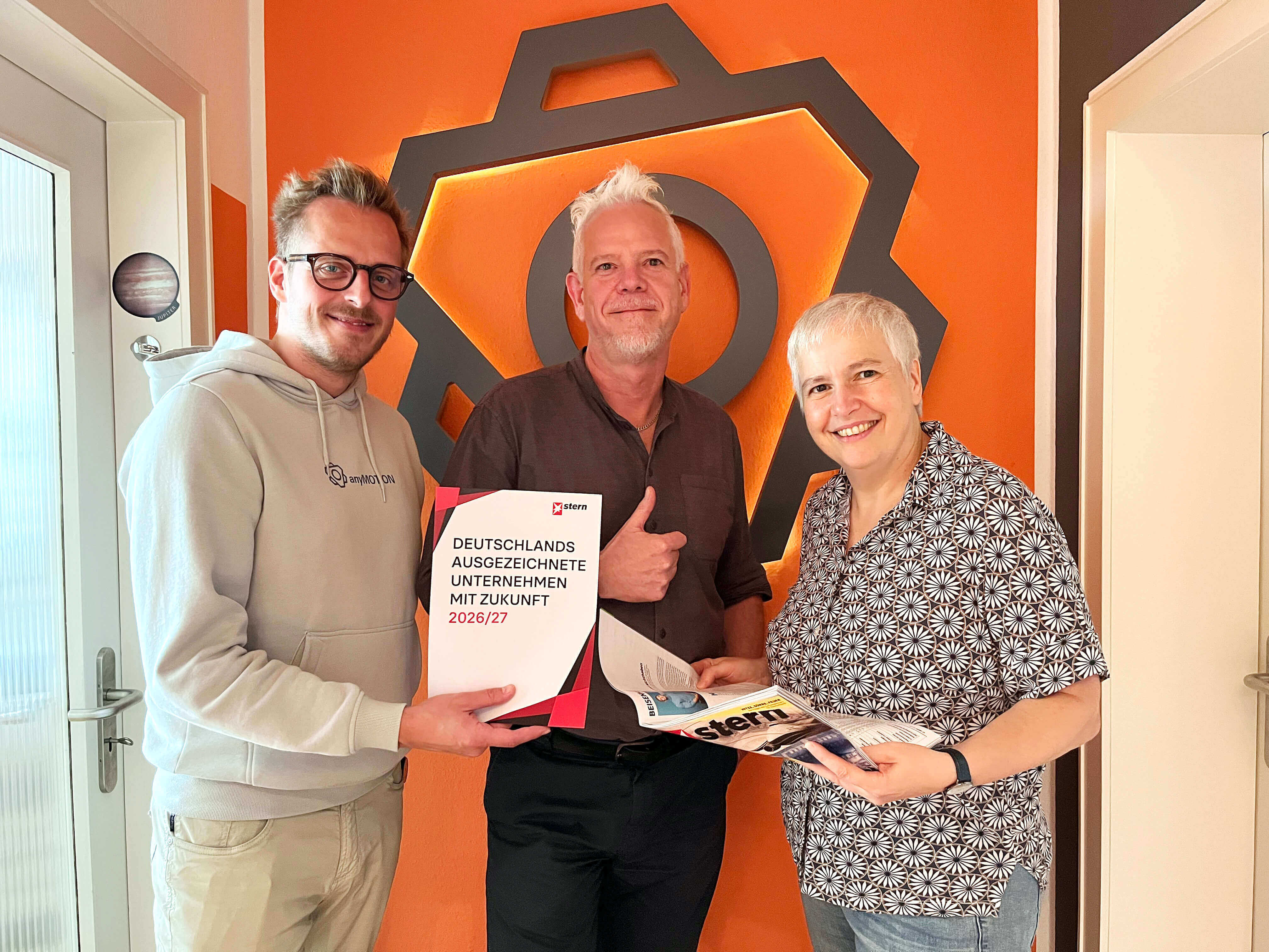 Drei Personen präsentieren eine Auszeichnung „Deutschlands ausgezeichnete Unternehmen mit Zukunft 2026/27“ vor einer orangefarbenen Wand mit anyMOTIONLogo.